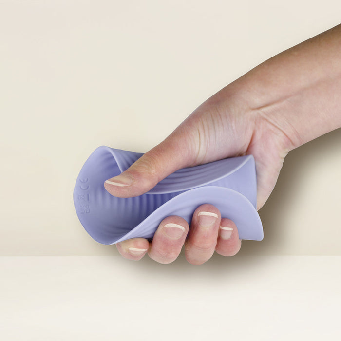 A person is squeezing the Niya N5 Flexible Lay On Humping Vibrator with Remote by Rocks Off in one hand. The background is a neutral off-white color. The lavender silicone vibrator features a ribbed texture and bends easily under the pressure of the hand.