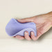 A person holds the Niya N5 Flexible Lay On Humping Vibrator with Remote in Lavender from Rocks Off. The light purple, wavy-textured silicone device is being gently squeezed, highlighting its soft and malleable nature. With waterproof features, it offers versatility in various settings. The background is a simple, light-colored surface.