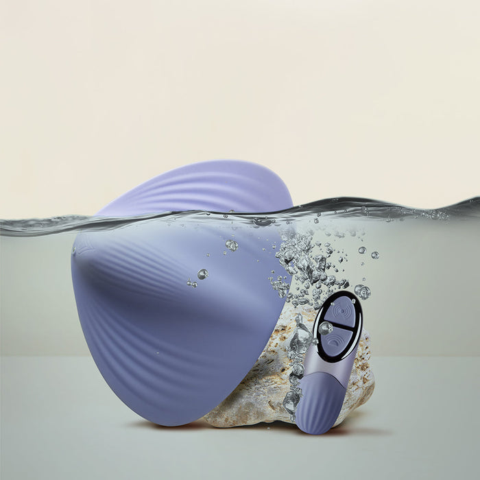 A lavender, clam-shaped Niya N5 Flexible Lay On Humping Vibrator with a ribbed texture, from Rocks Off, is partially submerged in water. Next to it sits a small cylindrical remote control with visible buttons. Water droplets splash around both waterproof items against a neutral background.