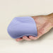 A hand is holding the Niya N5 Flexible Lay On Humping Vibrator with Remote in Lavender by Rocks Off, a light purple, flexible, ribbed device resembling a soft silicone pad. The background features neutral, light beige tones. The vibrator appears lightweight and pliable, perfectly shaped to fit comfortably in the palm.