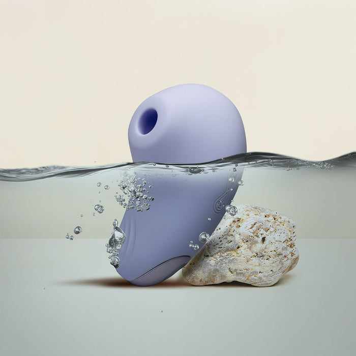 The image features the Niya N6 Ergonomic Easy Grip Clitoral Air Stimulator Vibrator by Rocks Off, a small, purple electronic device partly submerged in water and leaning against a textured rock. Bubbles around the device suggest movement in the water. Designed for clitoral suction, it promises orgasmic pleasure. The background transitions softly from cream to beige.