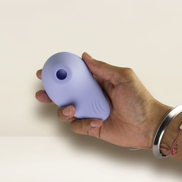 A hand, adorned with a silver bangle bracelet, holds the Niya N6 Ergonomic Easy Grip Clitoral Air Stimulator Vibrator by Rocks Off. The small, light purple, oval-shaped device features a circular indentation near the top for clitoral stimulation. Its smooth and ergonomic design is set against a plain, light-colored background.