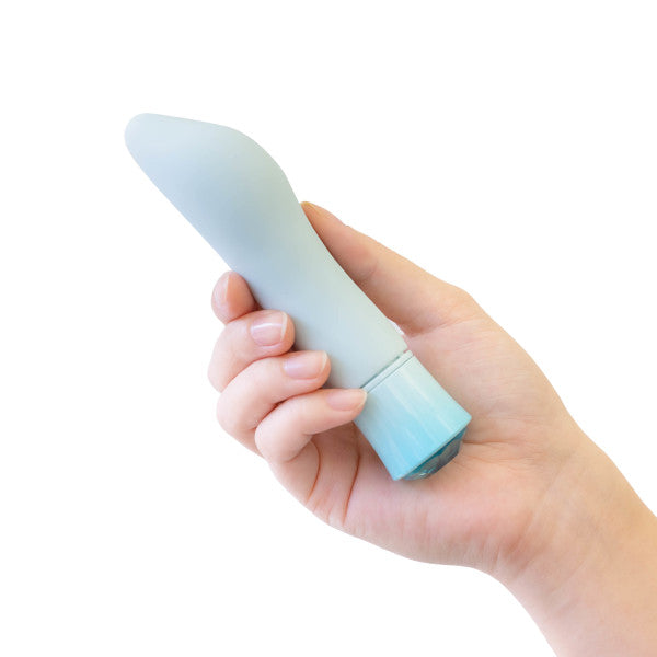 A hand holding the Oh My Gem Ardor Aquamarine Warming Internal & External Tapered Vibrator by Blush, showcasing its light blue, curved design with a rounded tip. The cylindrical vibrator has a slightly larger base in a varying blue hue. Its waterproof capabilities are emphasized against a white background, perfectly highlighting it in the hand.