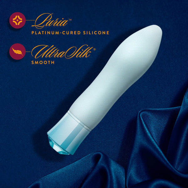 A white, curved vibrator with a blue base is placed on a blue silky fabric. Text beside it reads "Blush Oh My Gem Ardor Aquamarine, Platinum-Cured Silicone, UltraSilk Smooth." Two graphic logos accompany the text. Plus, it’s USB rechargeable for added convenience.