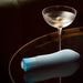 A refreshing martini with an olive sits on a dark, glossy table beside the sleek, light blue Oh My Gem Ardor Aquamarine Warming Internal & External Tapered Vibrator by Blush. The scene is set against a blurred background, highlighting the elegance of the drink and the contemporary design of the massager.