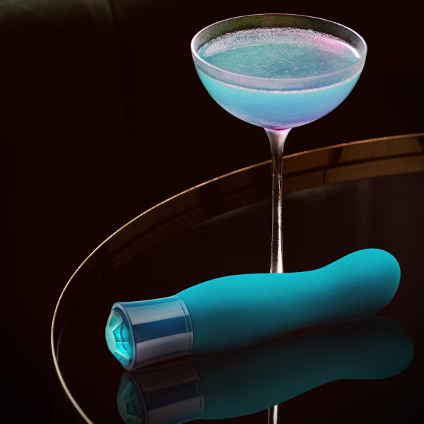 An Oh My Gem Fierce Blue Topaz Warming G-Spot Vibrator by Blush, meticulously crafted from phthalate-free silicone, lies on a reflective surface beside a cocktail glass filled with a shimmering blue and purple drink. The dimly lit background accentuates the vibrant colors of the objects.