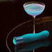 An Oh My Gem Fierce Blue Topaz Warming G-Spot Vibrator by Blush, meticulously crafted from phthalate-free silicone, lies on a reflective surface beside a cocktail glass filled with a shimmering blue and purple drink. The dimly lit background accentuates the vibrant colors of the objects.