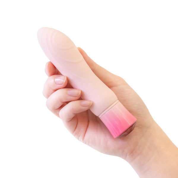 A hand holding the Blush Oh My Gem Morganite Warming Beginner Mini Vibrator in pale pink silicone with a darker pink base cap. The vibrator has a smooth, cylindrical shape with subtle ridges near the base and features multiple vibrating functions. The background is plain white.