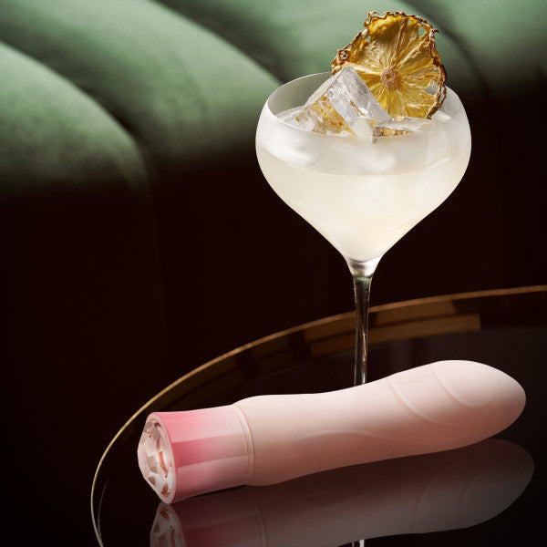 A stylish cocktail garnished with a dried pineapple slice sits on a sleek black surface. Next to the cocktail, there's an Oh My Gem Morganite Warming Beginner Mini Vibrator by Blush, featuring discreet vibrating functions in its chic pink and white design. A blurred green cushion appears in the background.