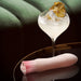 A stylish cocktail garnished with a dried pineapple slice sits on a sleek black surface. Next to the cocktail, there's an Oh My Gem Morganite Warming Beginner Mini Vibrator by Blush, featuring discreet vibrating functions in its chic pink and white design. A blurred green cushion appears in the background.