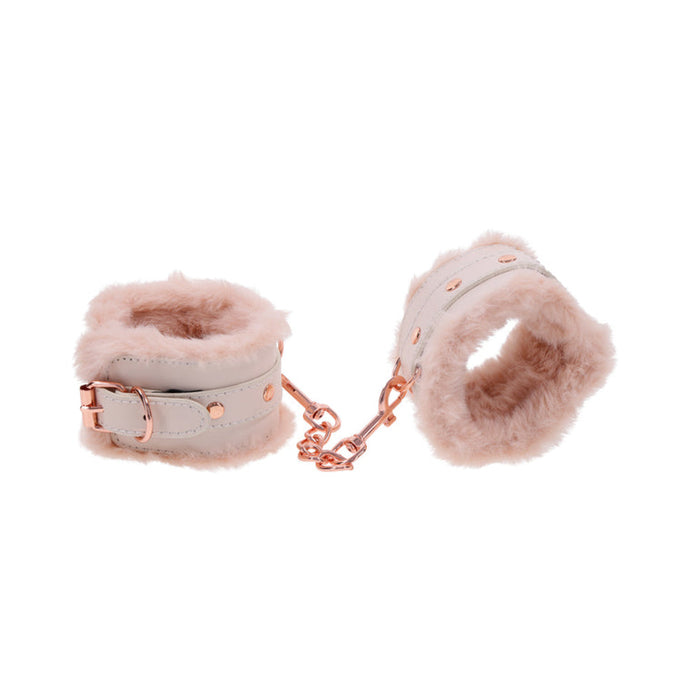 A pair of plush Peaches 'n Cream Fur Handcuffs by Sportsheets, featuring a soft pink faux fur design. These wrist cuffs are connected by a rose gold chain and adorned with rose gold buckles and small round studs, all showcased against a white background.