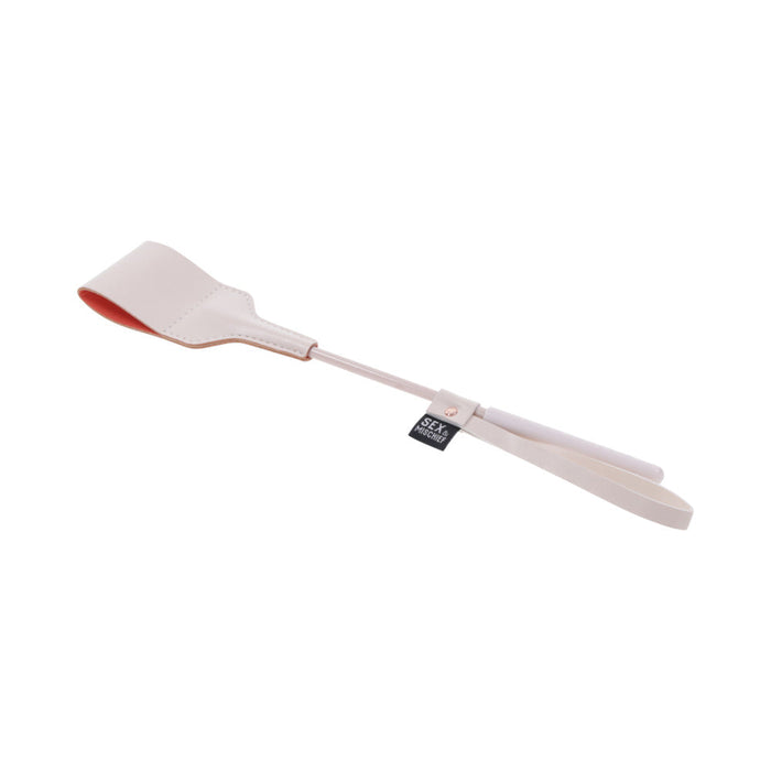 The Peaches 'n Cream Riding Crop by Sportsheets is crafted from cream-colored faux leather and features a rectangular flap at the end. It’s ideal for both equestrian pursuits and intimate moments, complete with a looped handle and a small black tag, all elegantly presented on a white background.