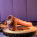 The Peek A Boo Uncut Vibrating 8" Silicone Caramel Dildo by Evolved Novelties sits elegantly on a wooden slab. The backdrop showcases a wooden panel wall adorned with scattered stones, all bathed in soft purple lighting that accentuates the sophisticated design.