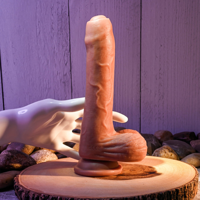 The Peek A Boo Uncut Vibrating 8" Silicone Caramel Dildo by Evolved Novelties stands upright on a wooden surface, surrounded by pebbles. It features a dual-layered and realistic design with a suction base. A white mannequin hand is positioned as if holding the dildo from behind, set against the backdrop of a textured wooden wall.
