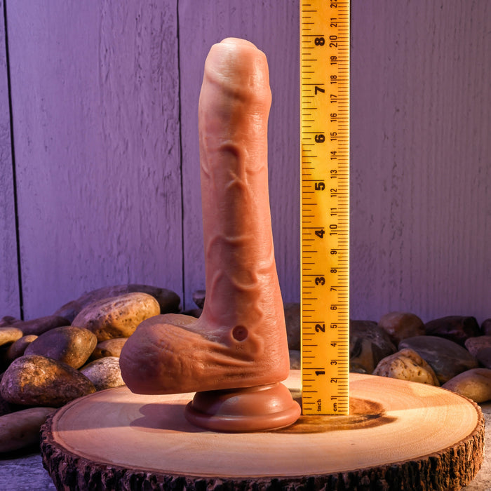 The Peek A Boo Uncut Vibrating 8" Silicone Caramel Dildo by Evolved Novelties stands upright on a wooden surface with a powerful suction cup base, next to a vertical measuring ruler indicating its height. The background features pebbles and a wooden textured wall.