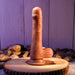 The Peek A Boo Uncut Vibrating 8" Silicone Caramel Dildo by Evolved Novelties, designed with dual-layered silicone and a powerful suction cup base, stands upright on a wooden surface. The background showcases wooden panels and smooth stones, creating a natural, rustic setting.