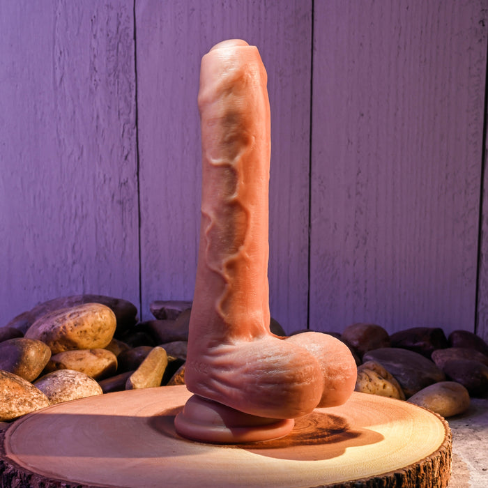 The Evolved Novelties Peek A Boo Uncut Vibrating 8" Silicone Caramel Dildo, featuring a realistic flesh-like texture and veins, stands vertically on a wooden surface surrounded by smooth stones. The backdrop showcases a light gray wooden panel softly lit with purple hues.