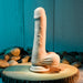 A beige Peek A Book Uncut Vibrating 8" Silicone Vanilla Dildo from Evolved Novelties, boasting detailed anatomical features, stands vertically on a round wooden base. Smooth river rocks are scattered around it, set against a rustic wooden background with a blue-green tint.