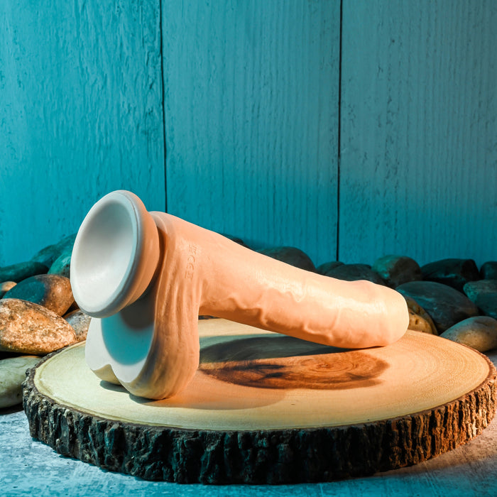 A 3D-printed bone model placed horizontally on a wooden surface, with a blue wooden plank background and smooth stones surrounding the base. The model, as anatomically detailed as the Peek A Book Uncut Vibrating 8" Silicone Vanilla Dildo by Evolved Novelties, is illuminated by soft light, casting shadows on the surface.