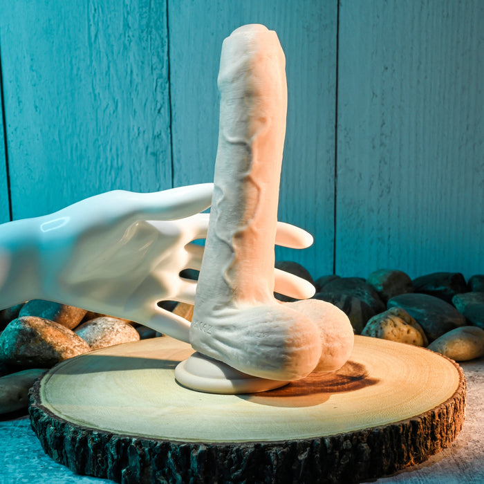 A white ceramic hand gracefully holds the Peek A Book Uncut Vibrating 8" Silicone Vanilla Dildo by Evolved Novelties, featuring a realistic dual-layered design with testicles. This elegant display is set on a circular wooden platform surrounded by smooth stones, against a backdrop of blue-tinted wooden texture.