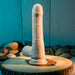 The Peek A Book Uncut Vibrating 8" Silicone Vanilla Dildo by Evolved Novelties stands upright on a wooden slab, surrounded by smooth stones. This realistic-looking, flesh-toned toy features two control buttons and detailed textures with veins. The background is a blue-toned wooden surface.