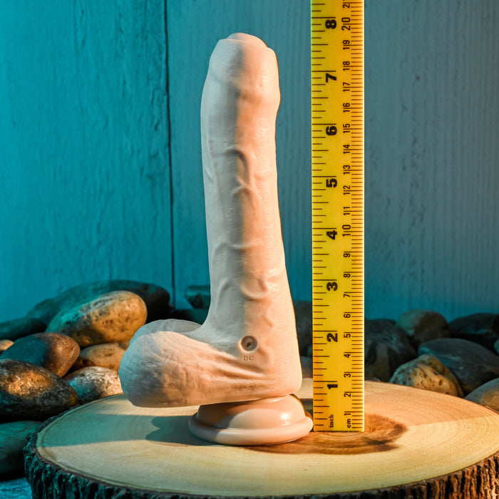 A Peek A Book Uncut Vibrating 8" Silicone Vanilla Dildo by Evolved Novelties stands upright on a round wooden base, placed on a surface surrounded by smooth stones. A yellow measuring tape stands beside it, showing the dildo's length as approximately 9 inches. The background is illuminated with teal lighting.