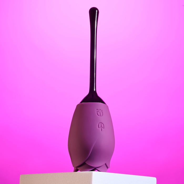 The Playboy Petal Flicking Tongue Double Ended Rose with Vibrating Stem by Evolved Novelties resembles a purple silicone flower bud with a long, sleek handle and sits on a white pedestal. Crafted from premium silicone, the background features a vibrant pink gradient, giving the image a modern and stylish look.