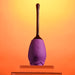 A sleek, purple Playboy Petal Flicking Tongue Double Ended Rose with Vibrating Stem from Evolved Novelties, designed to resemble a flower bud with a long, brown stem, is positioned upright on a clear stand against a gradient orange background. Crafted from premium silicone, the purple section features subtle floral imprint designs.