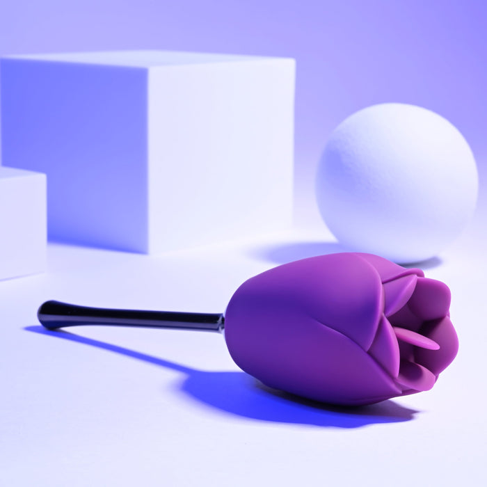 The Playboy Petal Flicking Tongue Double Ended Rose with Vibrating Stem from Evolved Novelties, a luxurious purple rose-shaped item crafted from premium silicone and featuring a black stem, rests on a surface against a backdrop of white geometric shapes. Soft blue-tinted lighting creates a calming and minimalistic scene.