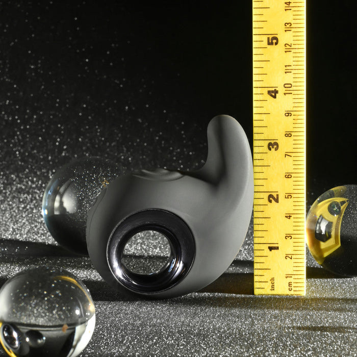 A smooth, black ergonomic object with a circular opening, placed on a glittery black surface, resembles the Playboy Ring My Bell Clitoral Thumper and Pinpoint Vibrator by Evolved Novelties. A yellow measuring tape stands beside it, showing a height of approximately 3 inches. Two glass spheres are positioned nearby, reflecting light.