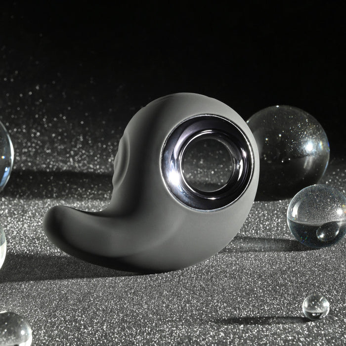 A sleek, black Playboy Ring My Bell Clitoral Thumper and Pinpoint Vibrator from Evolved Novelties, featuring a circular chrome accent, rests on a glittery, dark surface. Several clear, reflective spheres of varying sizes are scattered around it, creating a futuristic and abstract aesthetic. This USB rechargeable clitoral stimulator shines in the dark and sparkly background.