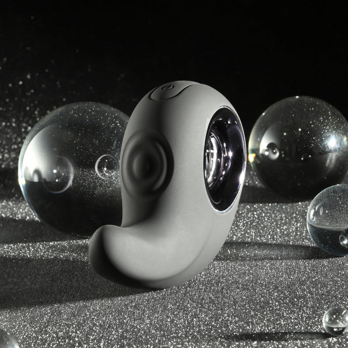 A sleek, black, curved device with a circular opening sits on a textured surface, surrounded by transparent glass orbs. The Playboy Ring My Bell Clitoral Thumper and Pinpoint Vibrator by Evolved Novelties is USB rechargeable and set against a dark, sparkling backdrop, giving the scene a futuristic vibe.