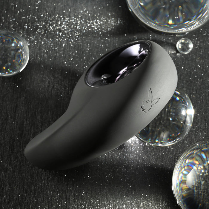 The Playboy Ring My Bell Clitoral Thumper and Pinpoint Vibrator - Black, designed by Evolved Novelties, rests on a dark surface amidst reflective glass spheres. This sleek, black ergonomic clitoral stimulator features a glossy interface with a small engraved bunny logo. Its modern design is aesthetically pleasing, accented by shimmering light reflections and is USB rechargeable.