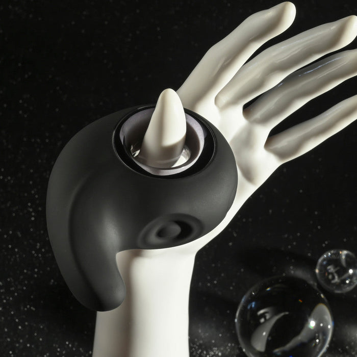 The Playboy Ring My Bell Clitoral Thumper and Pinpoint Vibrator by Evolved Novelties, featuring a sleek black finish and modern design, encircles a white mannequin hand with abstract, artistic lighting. The dark background is speckled with small, sparkling dots, evoking a starry night sky. Nearby, two transparent glass orbs add to the scene's artistic feel. This ergonomic device is USB rechargeable and waterproof.