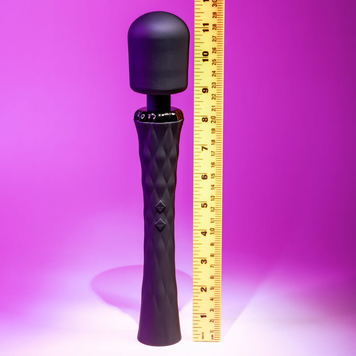 A Playboy Royal Rechargeable Black Silicone Wand Vibrator by Evolved Novelties with a slim handle stands upright on a purple background. A yellow measuring tape is placed beside it, displaying its height. The vibrator's textured handle features two buttons and a flexible neck for added comfort.