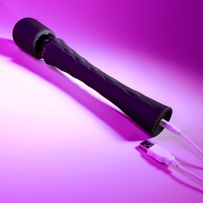 The Playboy Royal Rechargeable Black Silicone Wand Vibrator from Evolved Novelties, featuring a slim handle and flexible neck with a textured black finish, is displayed on a pink background. It is connected to a USB cable for charging.