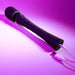 The Playboy Royal Rechargeable Black Silicone Wand Vibrator from Evolved Novelties, featuring a slim handle and flexible neck with a textured black finish, is displayed on a pink background. It is connected to a USB cable for charging.