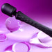 The Playboy Royal Rechargeable Black Silicone Wand Vibrator from Evolved Novelties, featuring a textured handle and two buttons, is laid on a pink and purple background. Transparent spherical and ring-shaped objects surround the wand, reflecting and refracting the colored light. Its flexible neck allows for better reach during use.