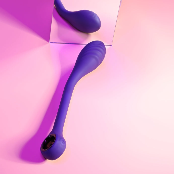 The Playboy Spot On Rechargeable Posable Silicone G-spot Vibrator from Evolved Novelties, an elongated, blue velvet-smooth silicone massager with a rounded handle, is positioned on a pink surface and angled towards a mirrored background that reflects the device. The scene is softly illuminated with pink and purple lighting, showcasing the sophisticated elegance and functional design of this USB rechargeable vibrator.