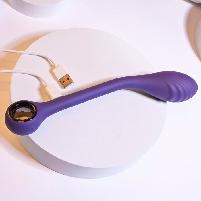 A purple Playboy Spot On Rechargeable Posable Silicone G-spot Vibrator by Evolved Novelties, featuring a curved design with a ring handle and ribbed tip, rests on a white circular platform. Crafted from velvet smooth silicone, it comes with USB rechargeability and is connected to a white charging cable.