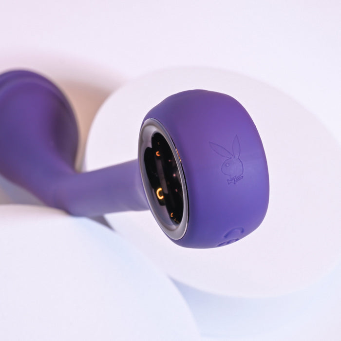 Close-up image of the Playboy Spot On Rechargeable Posable Silicone G-spot Vibrator by Evolved Novelties, featuring a purple design with a circular end and an engraved rabbit logo. The vibrator's velvet smooth silicone finish adds a luxurious touch, while the recessed area with gold contact points indicates USB rechargeable convenience. The background consists of white and purple surfaces.