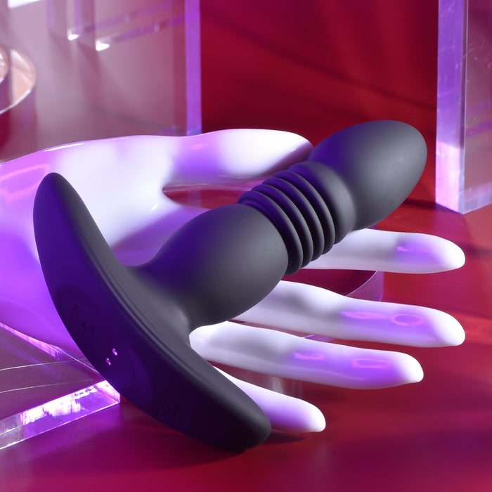 The Playboy Trust the Thrust Vibrating & Thrusting Anal Plug with Remote by Evolved Novelties, a sleek black ribbed butt plug with a flared base, is displayed in the grasp of a white mannequin hand. Boasting thrusting patterns and remote control capabilities, it stands against a backdrop of vibrant purple and red hues, crafting an energetic and contemporary aesthetic.