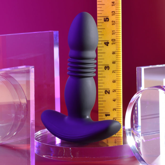 The Playboy Trust the Thrust Vibrating & Thrusting Anal Plug with Remote by Evolved Novelties, in sleek black silicone, stands vertically next to a yellow ruler indicating a height of approximately 4.5 inches. The background is illuminated with a pinkish glow and adorned with clear acrylic accessories, contributing to a contemporary aesthetic.