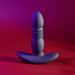 The Playboy Trust the Thrust Vibrating & Thrusting Anal Plug with Remote by Evolved Novelties, made of sculpted black silicone with a bulbous top, ribbed middle, and flared base, stands upright against a gradient background of dark red and purple.