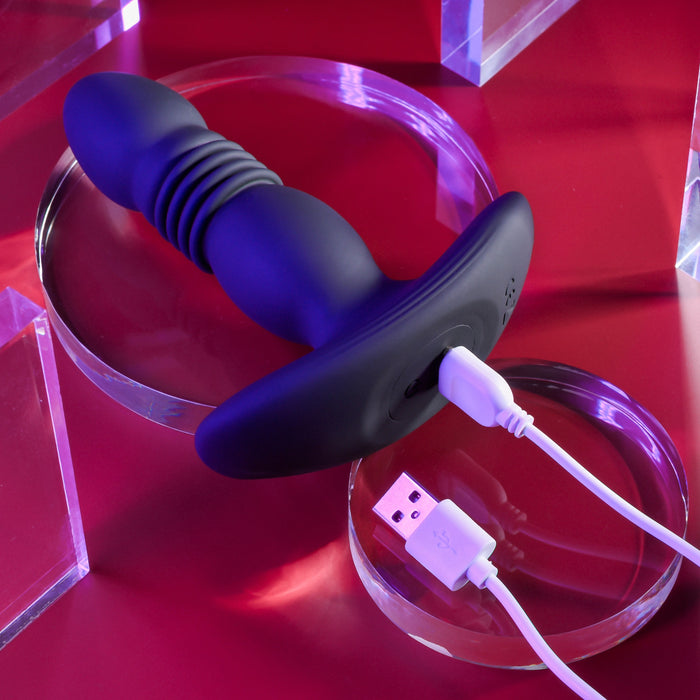 The Playboy Trust the Thrust Vibrating & Thrusting Anal Plug with Remote by Evolved Novelties, featuring a ribbed black silicone shaft and a flared base, is displayed on a red surface. A white USB charging cable is connected to its base. The toy rests on a transparent circular stand, surrounded by transparent rectangular blocks.