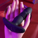 A Playboy Trust the Thrust Vibrating & Thrusting Anal Plug with Remote from Evolved Novelties, featuring a ribbed texture in the middle, rests on a mannequin hand with long, manicured nails. The scene is illuminated by purple and pink lighting, creating a sensual atmosphere.