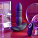 The Playboy Trust the Thrust Vibrating & Thrusting Anal Plug with Remote by Evolved Novelties, featuring a sleek dark surface, ribbed center design, and flared base, is displayed against a vibrant pink and red background. Accompanying it is a small remote control device. Surrounding the toy are various transparent and reflective objects creating an array of reflections.