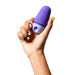 A hand with colorful, painted nails is holding a small, purple plastic device with two buttons on it. The WOW Romp Free X Beginner's Pleasure Air Clitoral Stimulator with Lid is smooth and has a slightly rounded shape. The background is plain white.