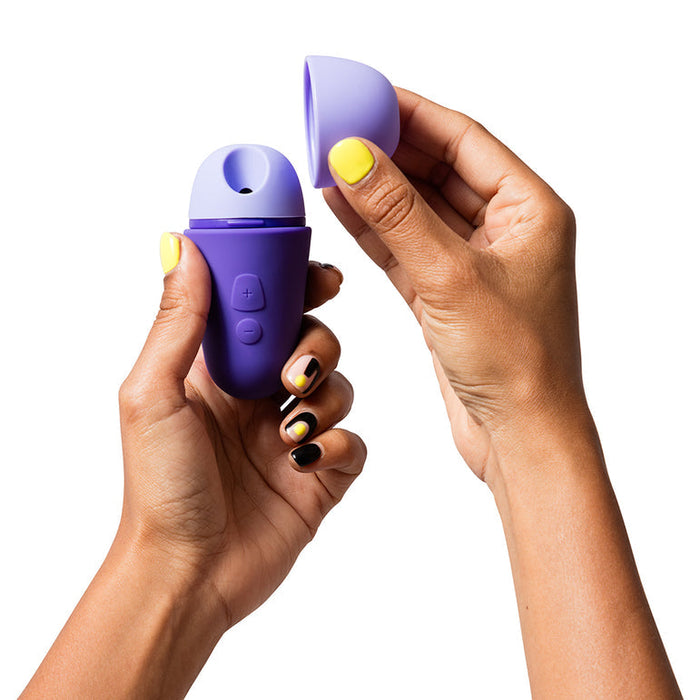 Hands with yellow and black nails hold the Romp Free X Beginner's Pleasure Air Clitoral Stimulator with Lid from WOW, a small purple cylindrical device. The cap is being removed to reveal a hole at the top, while plus and minus buttons are visible on the side of this travel-ready item. The background is white.