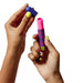 Two hands with colorful manicures hold a small, vibrant cylindrical object. The right hand grips a pink and blue patterned Romp Lipstick First Time Pleasure Air Clitoris Stimulator Vibrator from the brand WOW, while the left hand removes a blue cap. This device, featuring Pleasure Air Technology, promises heightened clitoral orgasms on a plain white background.